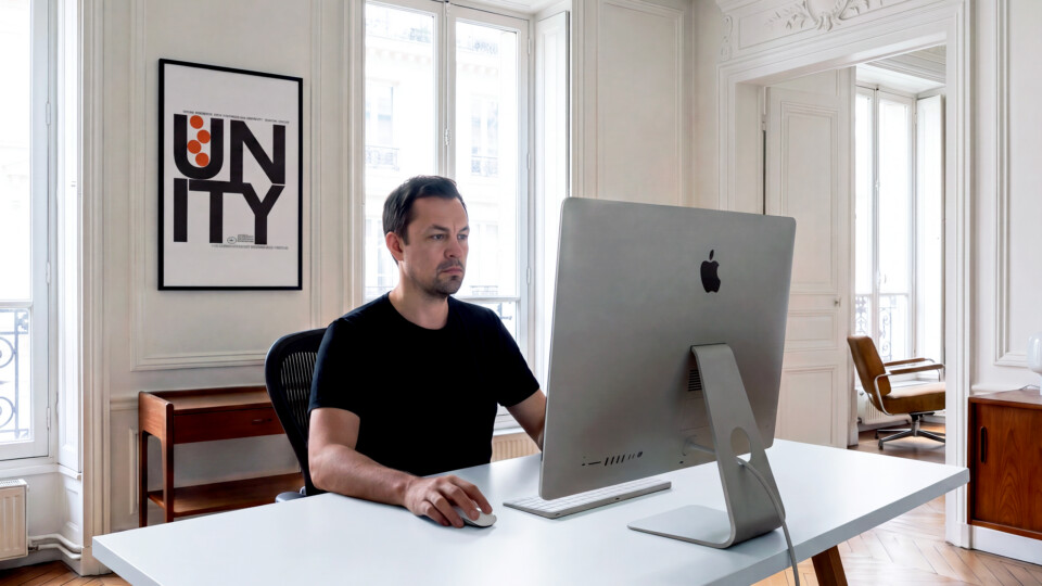 Jon Leverrier working at an iMac in a bright Haussmann-style apartment, with a framed typographic poster on the wall behind him and tall windows either side